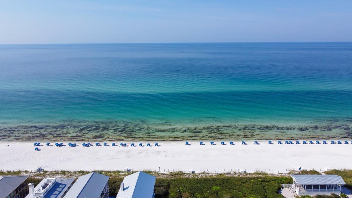 Aerial view of Scenic Highway 30A beachfront with white sand, turquoise Gulf waters, and rows of blue beach umbrellas near luxury vacation homes